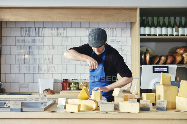 Cheesemonger cortar queso con alambre de queso rodeado de variedad de queso en la encimera - foto de stock