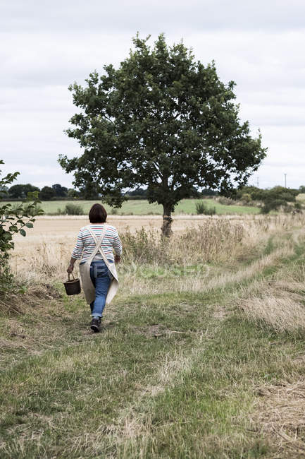 Rear view of woman walking along rural path and carrying brown wicker basket. — Stock Photo