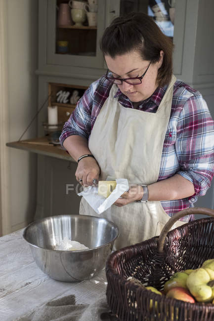 Femme dans le tablier debout dans la cuisine et couper des morceaux de beurre dans un bol en métal rempli de farine . — Photo de stock