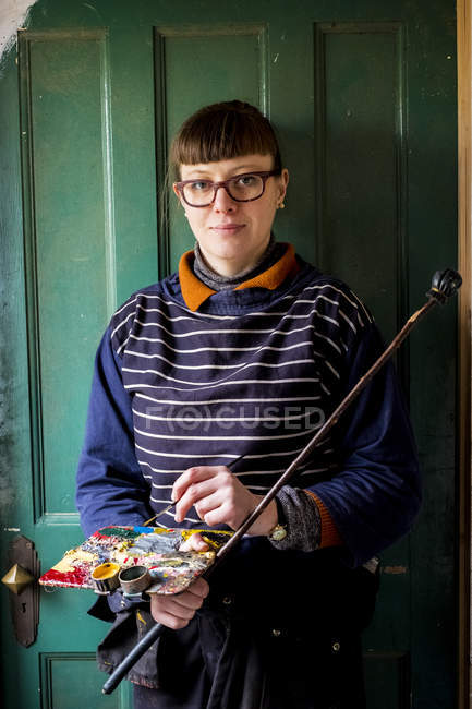 Woman in glasses holding paintbrush, palette and mahlstick while looking in camera. — Stock Photo