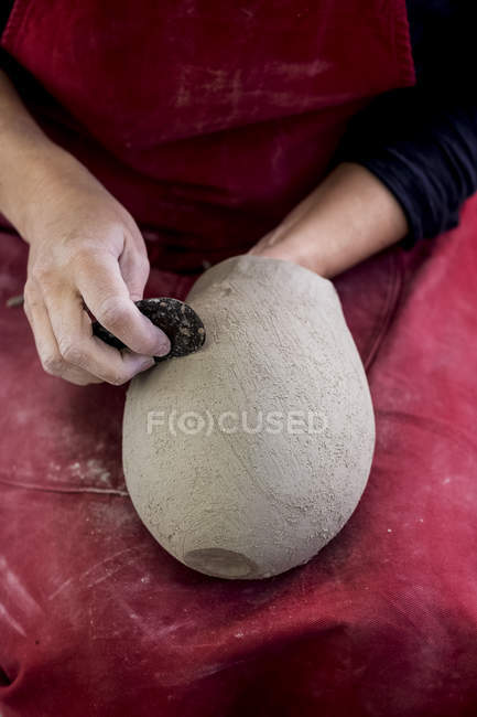 Sección media del artista de cerámica en delantal rojo sentado en el taller, trabajando en jarrón de arcilla
. - foto de stock