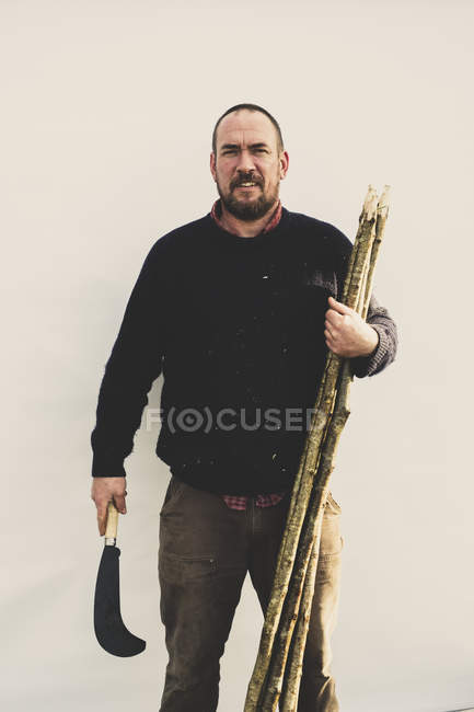 Hombre barbudo sosteniendo gancho de pico y montón de estacas de madera para la colocación de setos tradicionales, sonriendo en la cámara . - foto de stock