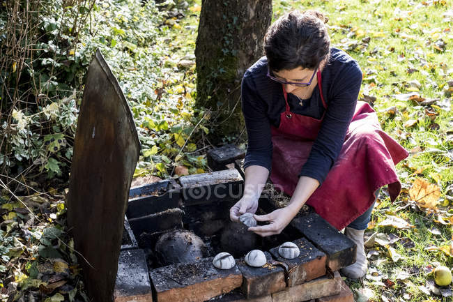 Female ceramic artist kneeling down at outdoor smoke fire pit and working on vase. — Stock Photo