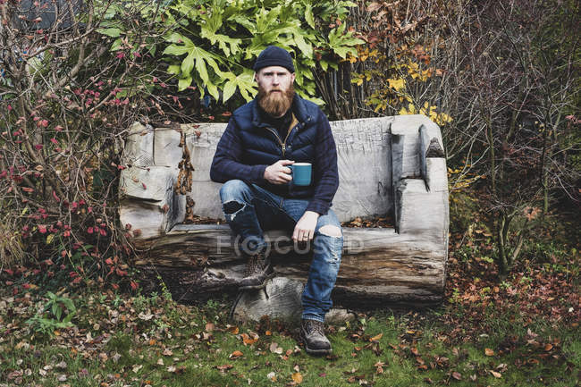Bearded uomo in berretto nero seduto su una panchina di legno in giardino, tenendo tazza blu, guardando in macchina fotografica
. — Foto stock