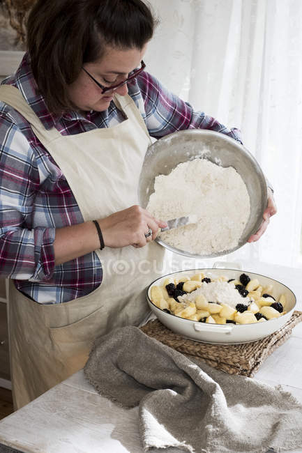 Woman wearing apron standing in kitchen, pouring fresh crumble mixture on pie dish filled with fruits. — Stock Photo