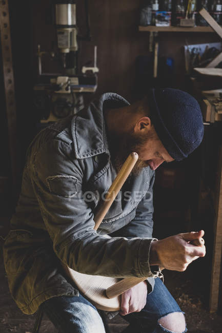 Bärtiger Mann mit schwarzer Mütze sitzt in Werkstatt und passt Bein an Holzhocker an. — Stockfoto