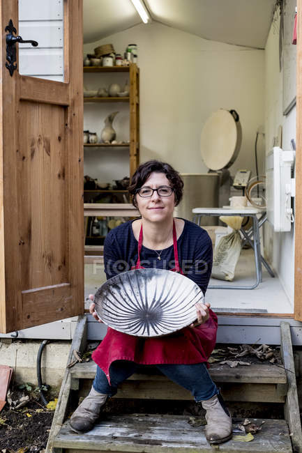 Woman in red apron sitting on steps outside pottery workshop and holding ceramic bowl with black lines pattern. — Stock Photo