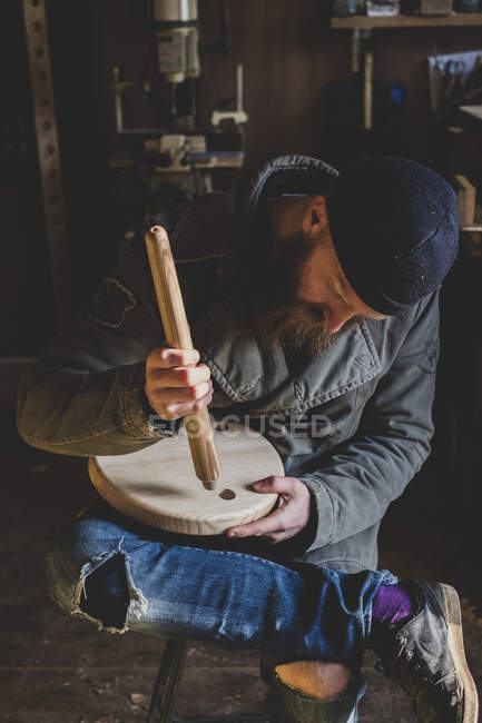 Bearded man in black beanie sitting in workshop, fitting leg to wooden stool. — Stock Photo