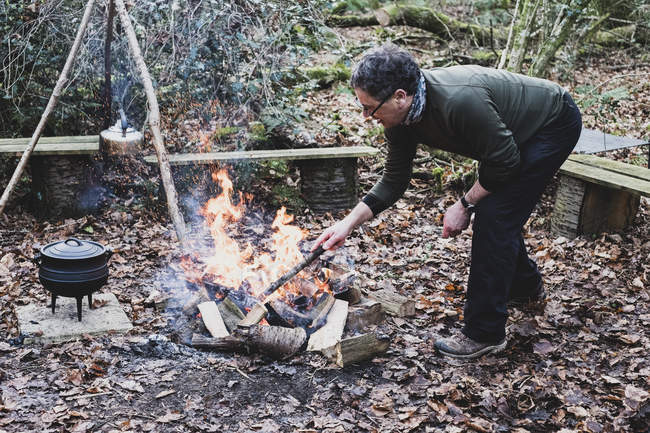 Hombre de pie en el bosque, avivando fogata con trípode de madera y olla de metal de hierro fundido
. - foto de stock