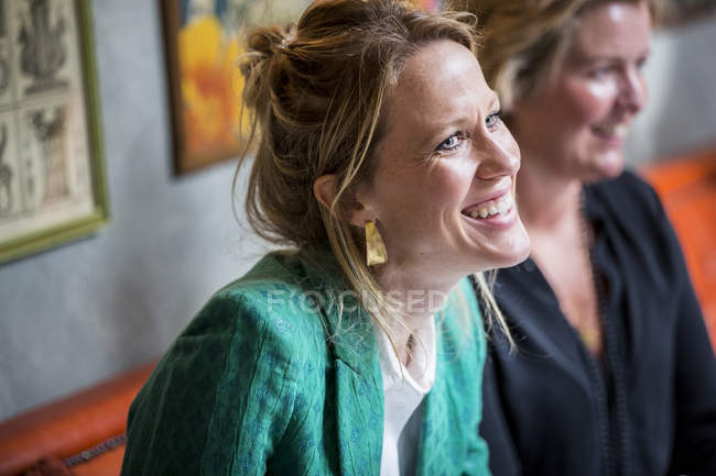 Mujer sonriente con cabello rubio en pendientes dorados y chaqueta verde sentada a la mesa . - foto de stock