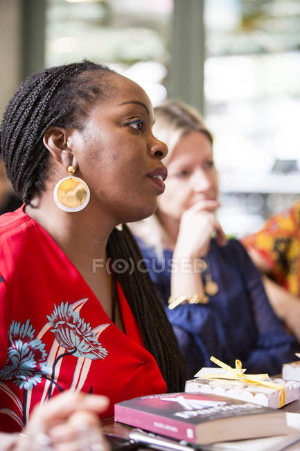 Mulher com cotovelos em brincos dourados e blusa vermelha sentada à mesa, ouvindo amigas atentamente . — Fotografia de Stock