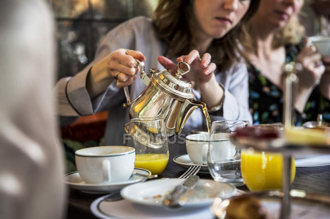 Середні дорослі жінки сидять за столом, поливаючи чай зі срібного чайного горщика . — стокове фото