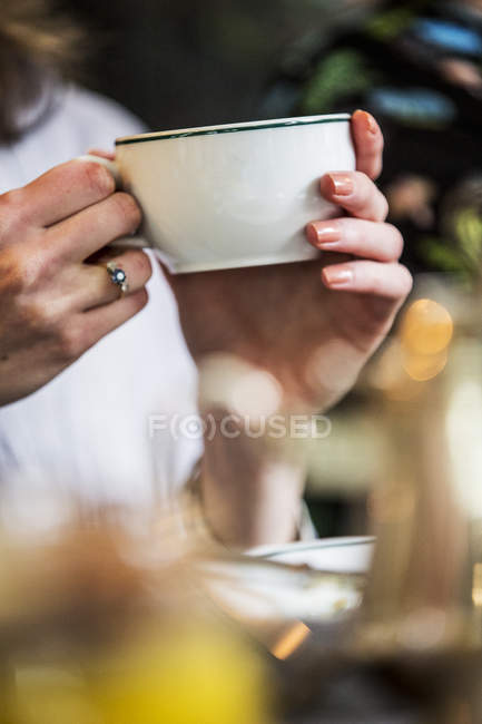 Close-up de mulher sentada à mesa e segurando copo de chá de porcelana branca . — Fotografia de Stock