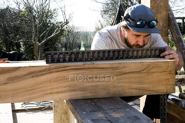 Man wearing baseball cap on building site, measuring wooden beam with metal ruler. — Stock Photo