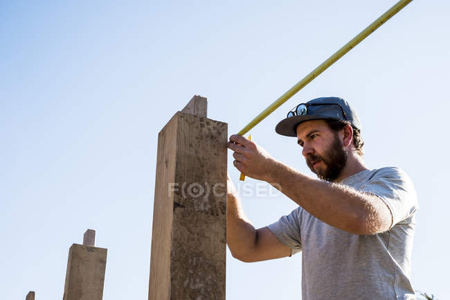 Man wearing baseball cap and sunglasses on building site, using tape measure. — Stock Photo