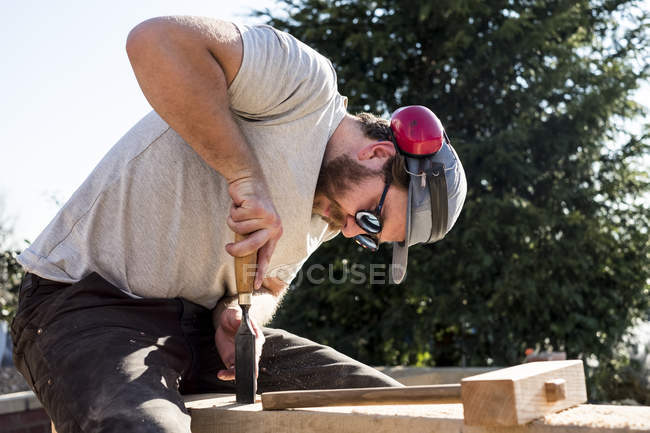 Homem usando boné de beisebol, óculos de sol e protetores auriculares no local de construção, trabalhando em feixe de madeira . — Fotografia de Stock