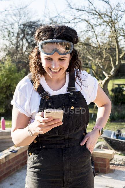 Smiling woman wearing dungarees and protective goggles checking mobile phone. — Stock Photo