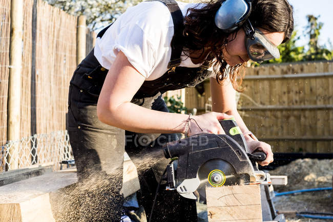 Frau mit Schutzbrille und Ohrenschutz hält Kreissäge in der Hand, schneidet Stück Holz auf Bauseite. — Stockfoto