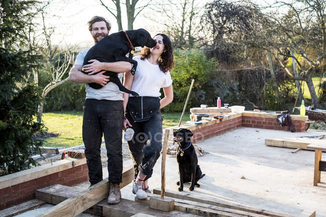 Smiling man and woman and two black dogs on building site of residential building. — Stock Photo