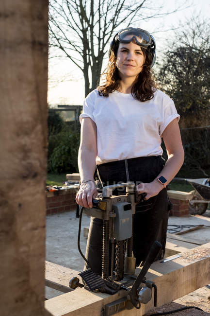 Woman wearing protective goggles holding machine, working on wooden building frame. — Stock Photo