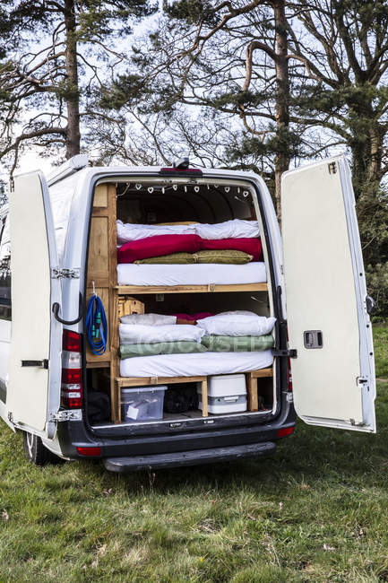 Vista trasera de la caravana estacionada en el prado con pilas de colchones y ropa de cama en el vehículo . - foto de stock