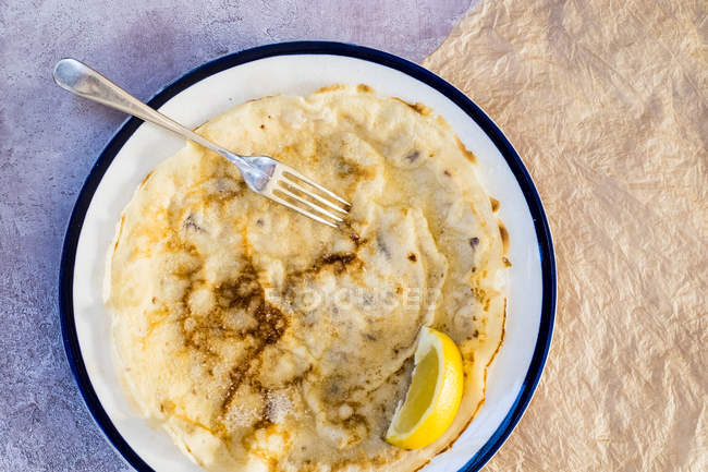 Nahaufnahme von frisch gebackenem Crêpe mit Zitronenkeil garniert auf weißem Teller mit blauem Rand. — Stockfoto
