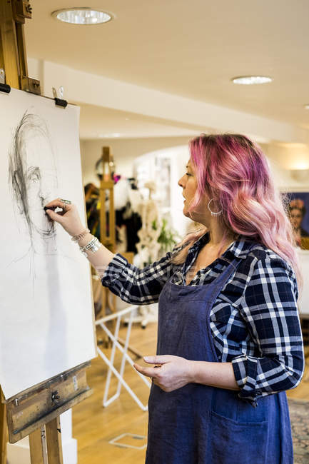 Женщина с длинными светлыми волнистыми волосами с розовыми полосками в фартуке, стоящая у мольберта и рисующая портрет . — стоковое фото