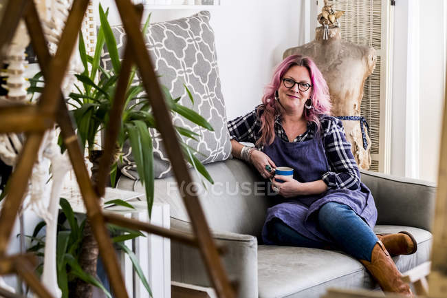 Mujer sonriente con el pelo largo y rubio ondulado con rayas rosadas sentado en el sofá y mirando en la cámara . - foto de stock