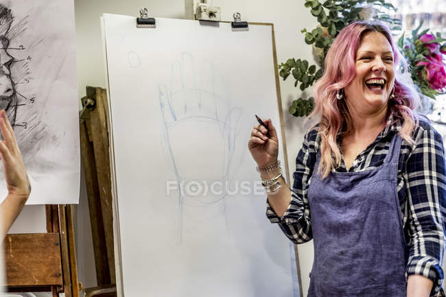 Laughing woman in apron standing at easel and drawing of hand. — Stock Photo