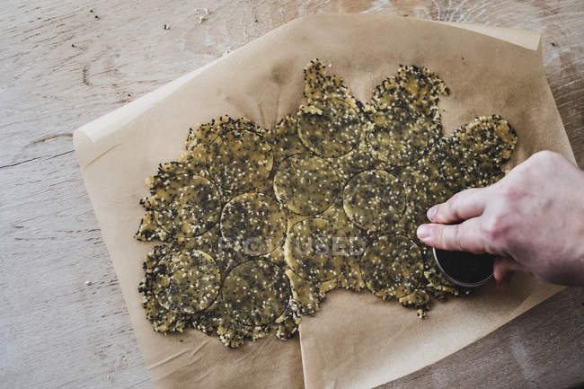 High angle close-up of person hand cutting discs of seeded cracker dough. — Stock Photo