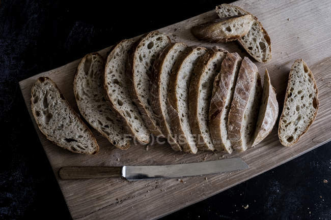 Vista en ángulo alto de la hogaza de pan marrón fresco en rodajas sobre tabla de madera con cuchillo afilado - foto de stock