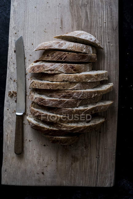 Vista en ángulo alto de la hogaza de pan marrón fresco en rodajas sobre tabla de madera con cuchillo afilado - foto de stock