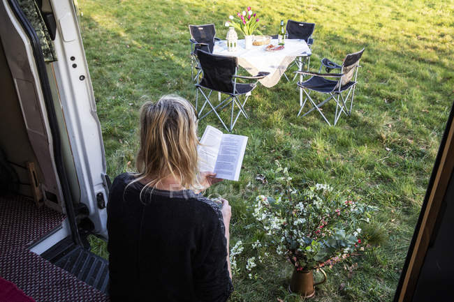 Vista trasera de la mujer sentada en la puerta de la caravana estacionada en el prado y libro de lectura . - foto de stock