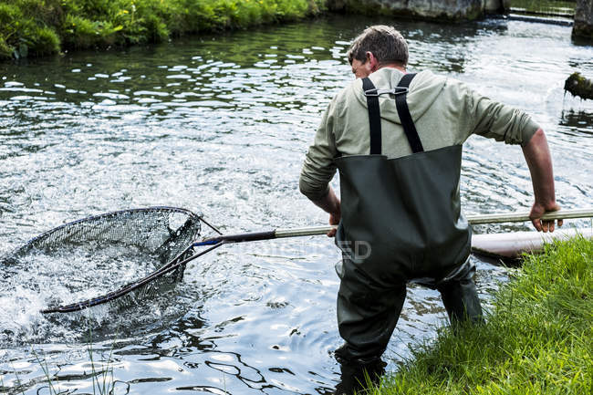 Mann in Watstiefeln steht im Flusswasser und hält großes Fischnetz in der Hand. — Stockfoto