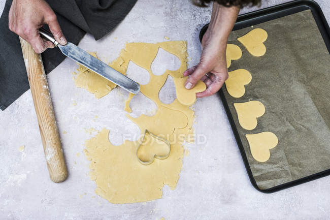 Primer plano de alto ángulo del cocinero utilizando un cuchillo de paleta para mover galletas en forma de corazón en la bandeja de hornear . - foto de stock