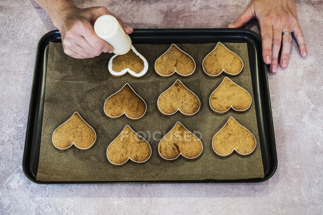High angle close-up of male cook decorating freshly baked heart-shaped cookies with piped white icing. — Stock Photo