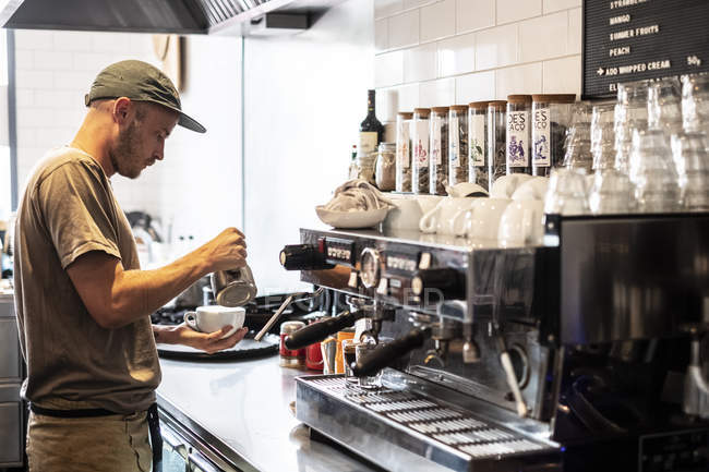 Bärtiger Mann mit Baseballmütze steht und kocht Kaffee an Espressomaschine eines Restaurants. — Stockfoto