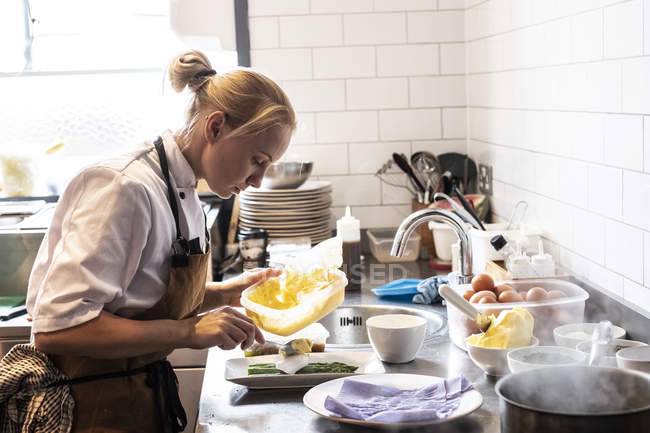 Жіночий шеф-кухарка в коричневому фартусі стоїть за кухонною стійкою, прикрашає тарілку спаржі . — стокове фото