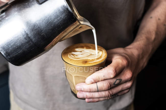 Close-up de pessoa com dedos tatuados derramando leite de jarro de metal em vidro de café latte . — Fotografia de Stock
