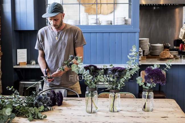 Homem em boné de beisebol em pé à mesa, cortando cardos para arranjos de flores em frascos de vidro grandes . — Fotografia de Stock