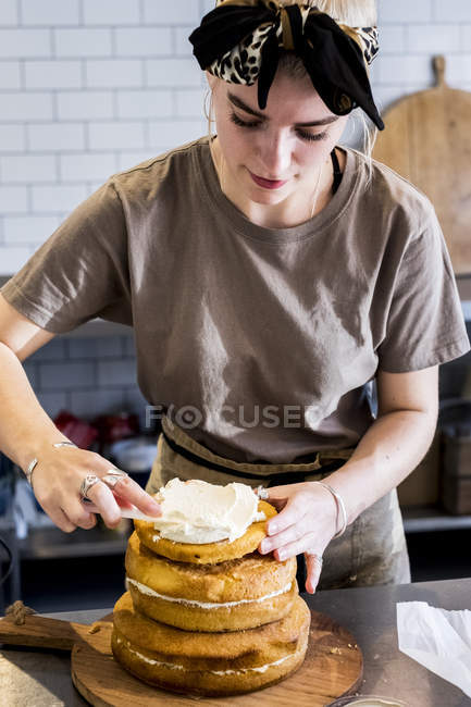 Cocinera femenina trabajando en cocina comercial montando torta de esponja en capas con crema fresca . - foto de stock