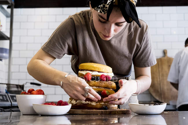 Female cook working in commercial kitchen assembling layered sponge cake with fresh fruits. — Stock Photo