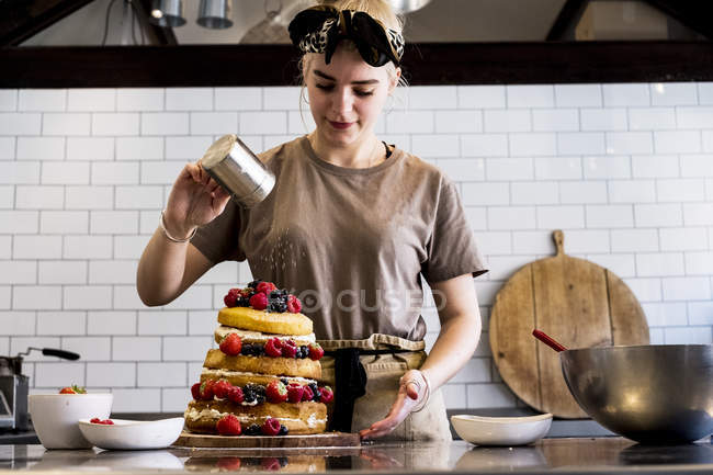 Cozinheiro feminino trabalhando na cozinha comercial polvilhando açúcar de confeiteiro sobre bolo em camadas com frutas frescas . — Fotografia de Stock