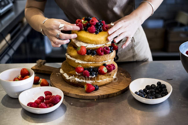 Midsection of cook working in commercial kitchen arranging fresh fruits over layered cake with fresh cream. — Stock Photo