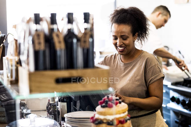 Mulher de raça mista trabalhando no café, pilha de pratos, bolo de esponja em camadas com creme fresco e frutas frescas em primeiro plano . — Fotografia de Stock