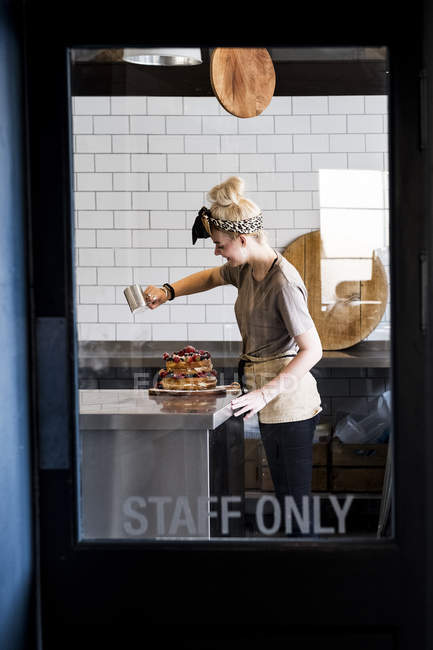 View through glass door marked Staff Only, of female cook working in commercial kitchen sprinkling icing sugar over layered cake with fresh fruits. — Stock Photo