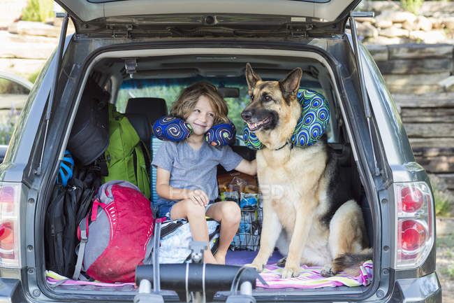 Menino idade elementar com cão pastor alemão vestindo travesseiros de viagem na parte de trás do SUV . — Fotografia de Stock