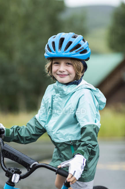 Портрет хлопчика початкового віку в дощовій куртці та шоломі, що розтягує велосипед . — стокове фото