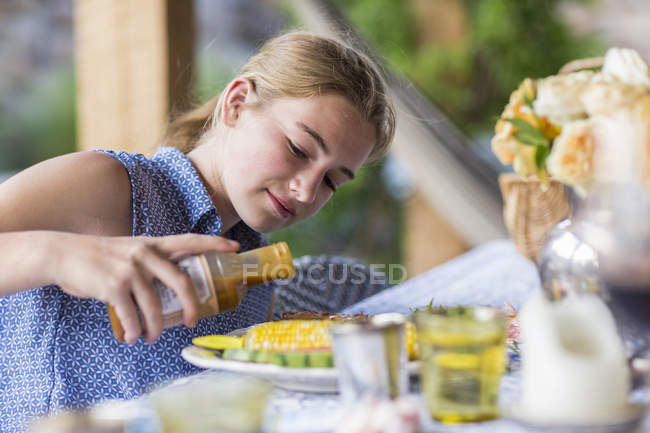 Blonde teenage girl pouring BBQ sauce on ribs and corn. — Stock Photo