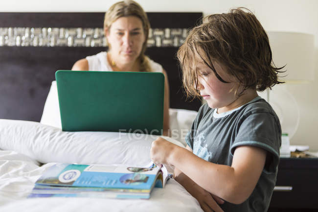 Boy reading book in hotel room as mother working on laptop in bed. — Stock Photo